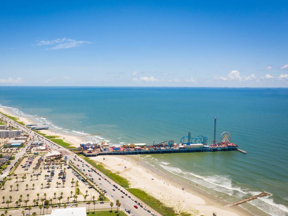 Aerial view of Galveston. From left to right are rows of homes, beach, Pleasure Pier, and bright blue waters