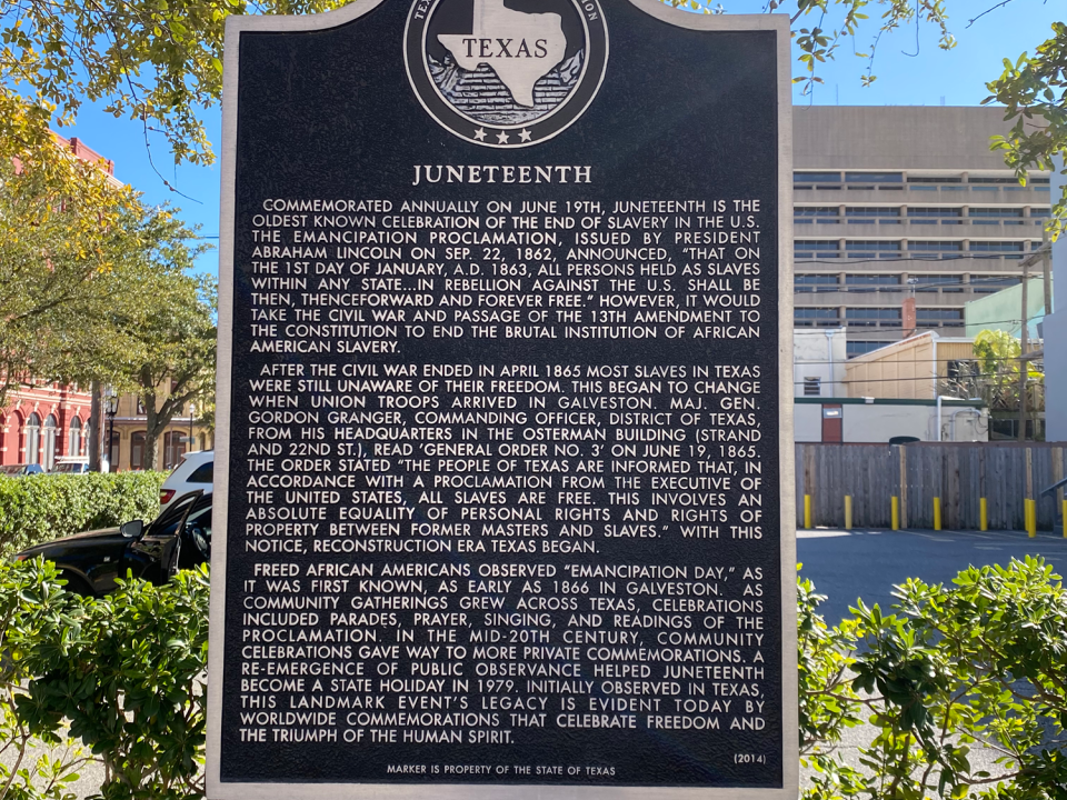 Juneteenth historical marker found in Galveston Texas. The marker is surrounded by greenery and buildings are in the distance