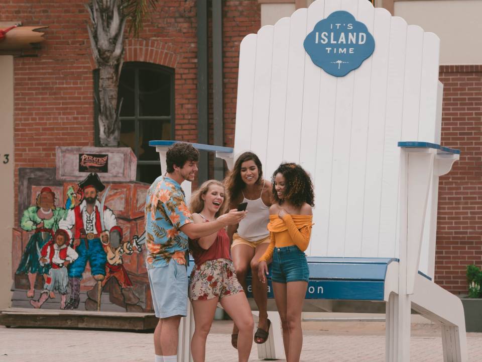 group of friends smiling at phone while standing in front of an oversized beach chair that says "It's Island Time"